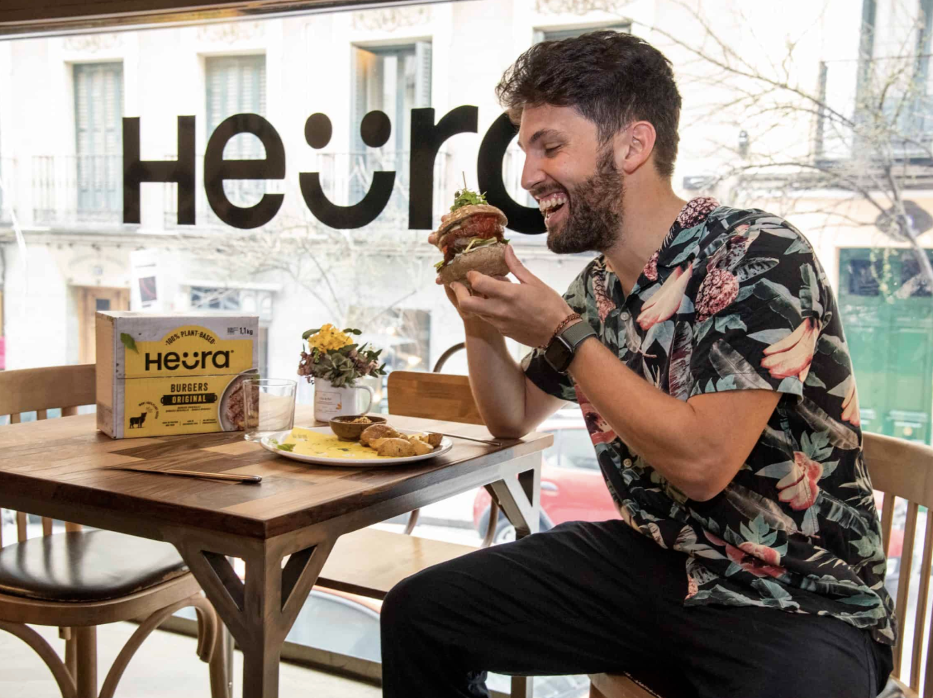 bearded person seated at a table about to bite into a plant-based burger. Heura box is on the table.